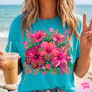 May include: Turquoise t-shirt featuring a vibrant floral design. The design includes pink hibiscus flowers with leopard print centers, surrounded by green leaves and smaller pink flowers. The shirt has short sleeves and a crew neck.