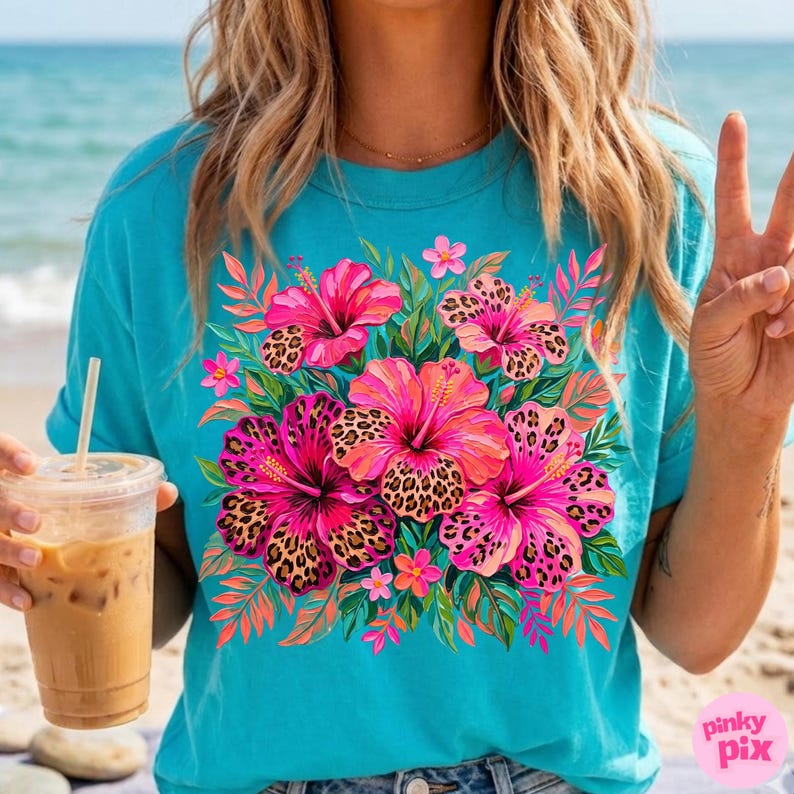 May include: Turquoise t-shirt featuring a vibrant floral design. The design includes pink hibiscus flowers with leopard print centers, surrounded by green leaves and smaller pink flowers. The shirt has short sleeves and a crew neck.