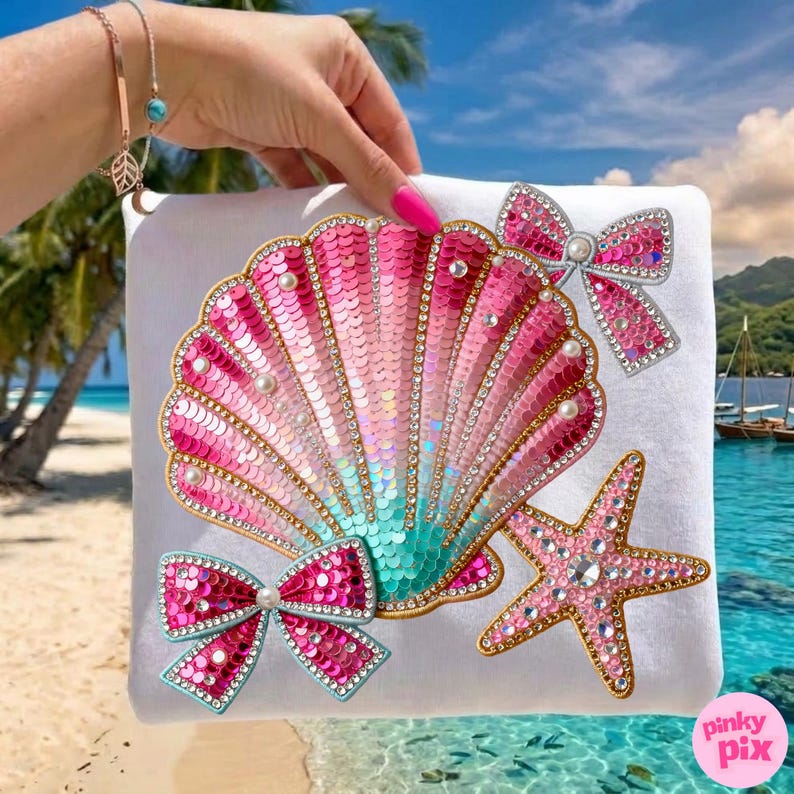 May include: White square pillow with a sequined seashell design in shades of pink and turquoise, adorned with pearls and rhinestones. Features a starfish and two bow accents, all embellished with sequins and rhinestones. The pillow has the text "pinky pix" in the bottom right corner.