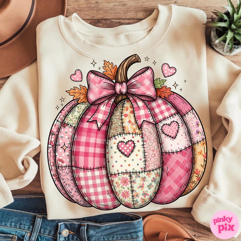May include: Cream-colored sweatshirt featuring a patchwork pumpkin design. The pumpkin is composed of various pink patterns, including gingham, floral, and polka dots, with a large pink bow and heart accents. The design has a fall theme.