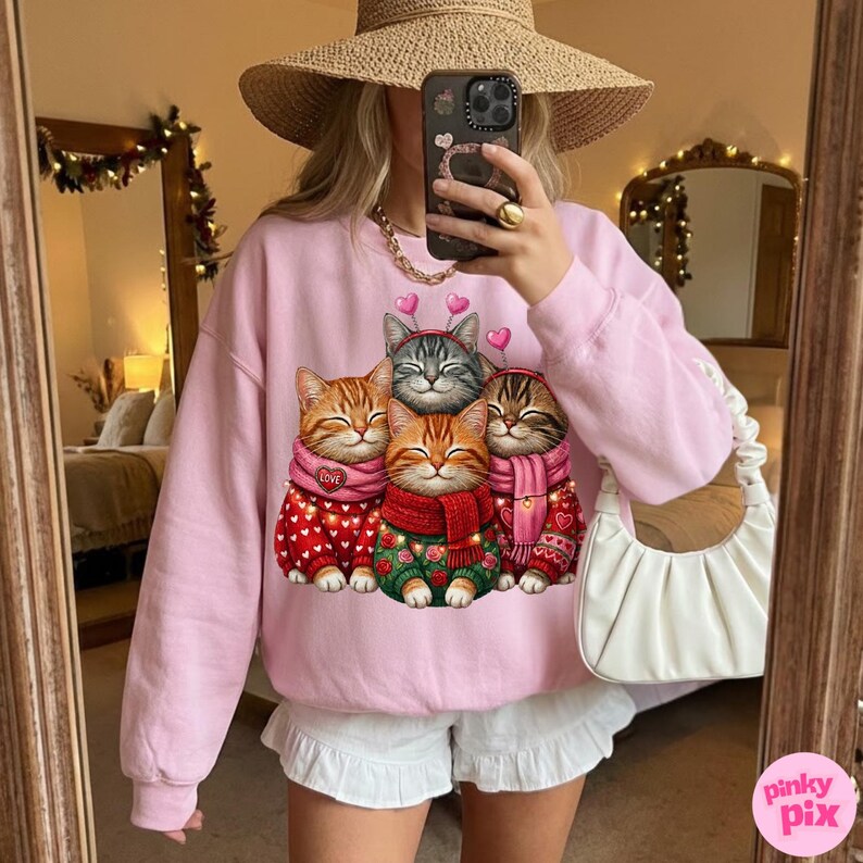 May include: A light pink sweatshirt featuring a graphic of four cartoon cats wearing scarves and sweaters. The cats are in shades of orange, gray, and brown. The sweatshirt has a crew neck and long sleeves.