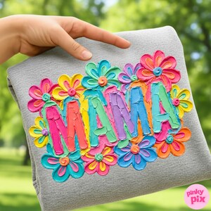 May include: A gray sweatshirt with the word "MAMA" in colorful, textured lettering, surrounded by vibrant, layered flower designs in shades of pink, yellow, blue, and orange. The design is held up by a hand.