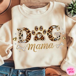 May include: Cream-colored sweatshirt with the text "DOG Mama" in a mix of leopard print and rhinestone embellishments. The design includes a paw print with a heart and bone accents, perfect for dog lovers.