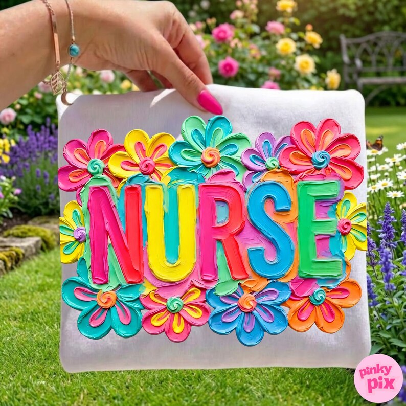 May include: A white square pillow with the word "NURSE" in vibrant, raised lettering, surrounded by colorful, textured flowers. The flowers feature shades of pink, yellow, blue, and orange. The pillow is held against a backdrop of green grass and flowers.