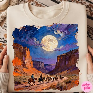 May include: Cream-colored sweatshirt featuring a vibrant, textured painting of a desert canyon scene under a full moon. Cowboys on horseback ride along a path. The artwork uses shades of blue, orange, and purple.