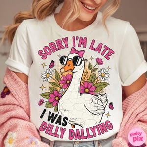 K&ouml;nnte beinhalten: Wei&szlig;es T-Shirt mit einer Grafik einer Gans mit Sonnenbrille und rosa Schleife. Der Text lautet "Sorry I'm Late, I Was Dilly Dallying" in Pink. Das Design umfasst Blumen und Schmetterlinge.