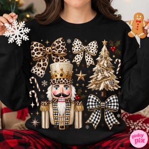 May include: A black sweatshirt featuring a central nutcracker graphic with a leopard print crown and uniform details. The design includes a Christmas tree, candy canes, and three bows in leopard, polka dot, and black and white buffalo check patterns.