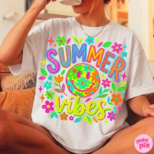 May include: White t-shirt with a colorful "SUMMER VIBES" graphic. The design features a smiley face with flower details, surrounded by flowers and leaves in bright neon colors. The shirt is a casual, relaxed fit.