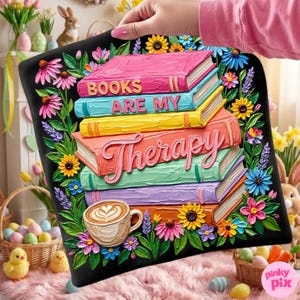 May include: A black decorative pillow featuring a stack of colorful books with the words "BOOKS ARE MY Therapy". The pillow is adorned with flowers and a coffee cup. The image is held by a person.
