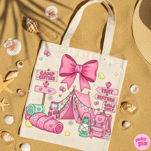 May include: Cream tote bag featuring a pink bow and camping illustrations. The design includes a tent, sleeping bag, backpack, and the text "Camp Cuties" and "Tent Sisters Girls Only". The bag is surrounded by seashells on a sandy surface.