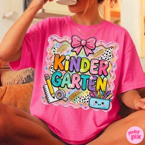 May include: A bright pink t-shirt with a colorful "Kindergarten" graphic. The design includes a pink bow, pencils, a pencil case, and a floral border. The shirt is a comfortable, casual style.