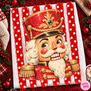 May include: A white fabric item featuring a red and white polka dot striped background with a central illustration of a nutcracker wearing a red uniform, gold epaulettes, and a tall hat with a green jewel accent and white hair.