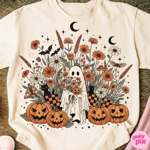 May include: Cream-coloured t-shirt with a Halloween graphic. The design features a ghost holding a pumpkin, surrounded by orange and white flowers, black cats, bats, and jack-o'-lanterns. Two crescent moons and stars complete the design.