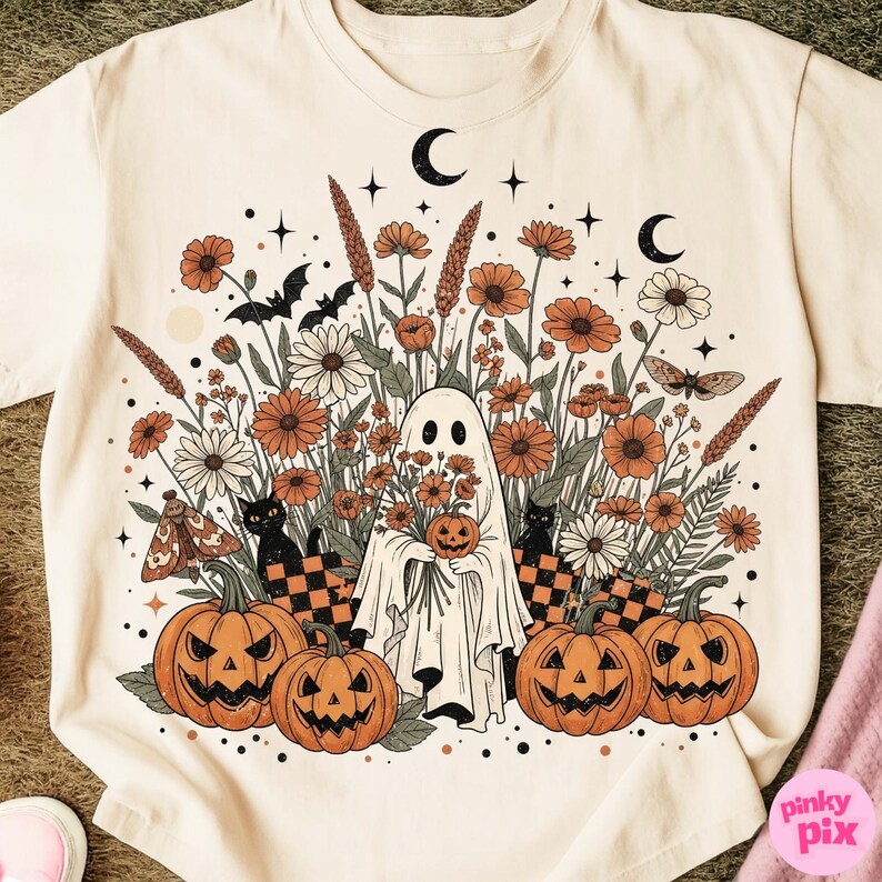 May include: Cream-coloured t-shirt with a Halloween graphic. The design features a ghost holding a pumpkin, surrounded by orange and white flowers, black cats, bats, and jack-o'-lanterns. Two crescent moons and stars complete the design.