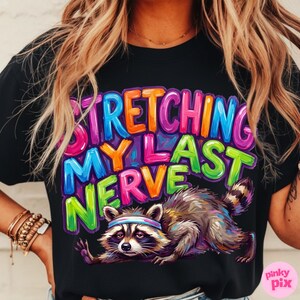 May include: Black t-shirt with the phrase "STRETCHING MY LAST NERVE" in colorful, bubble letters. A raccoon wearing a headband is stretching below the text. The shirt has a relaxed fit.