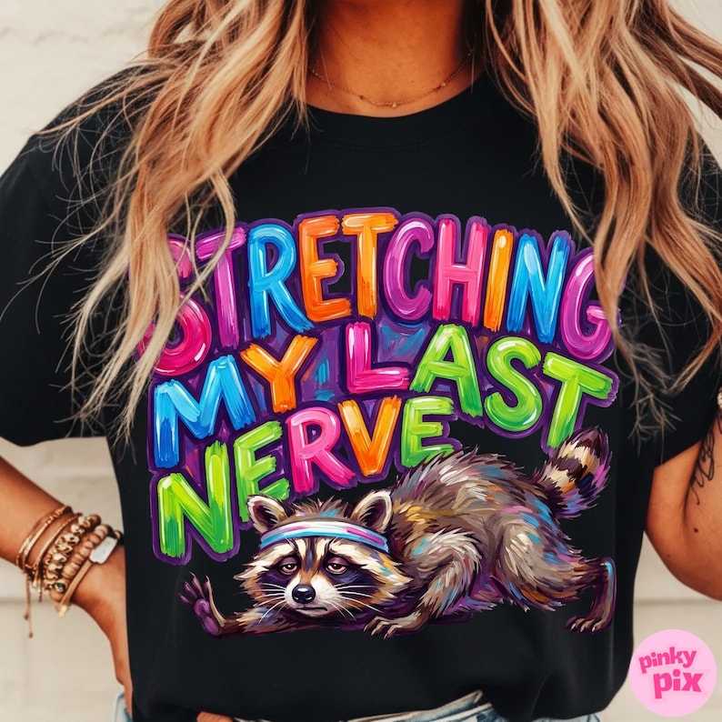 May include: Black t-shirt with the phrase "STRETCHING MY LAST NERVE" in colorful, bubble letters. A raccoon wearing a headband is stretching below the text. The shirt has a relaxed fit.