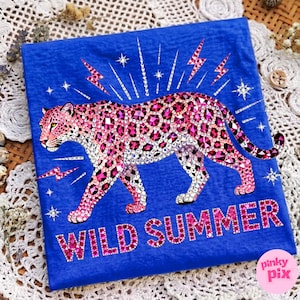 May include: A blue t-shirt featuring a pink leopard design with rhinestone accents. The leopard is surrounded by lightning bolts and stars. The words "WILD SUMMER" are displayed in pink rhinestones. The shirt has a Pinky Pix logo in the bottom right corner.