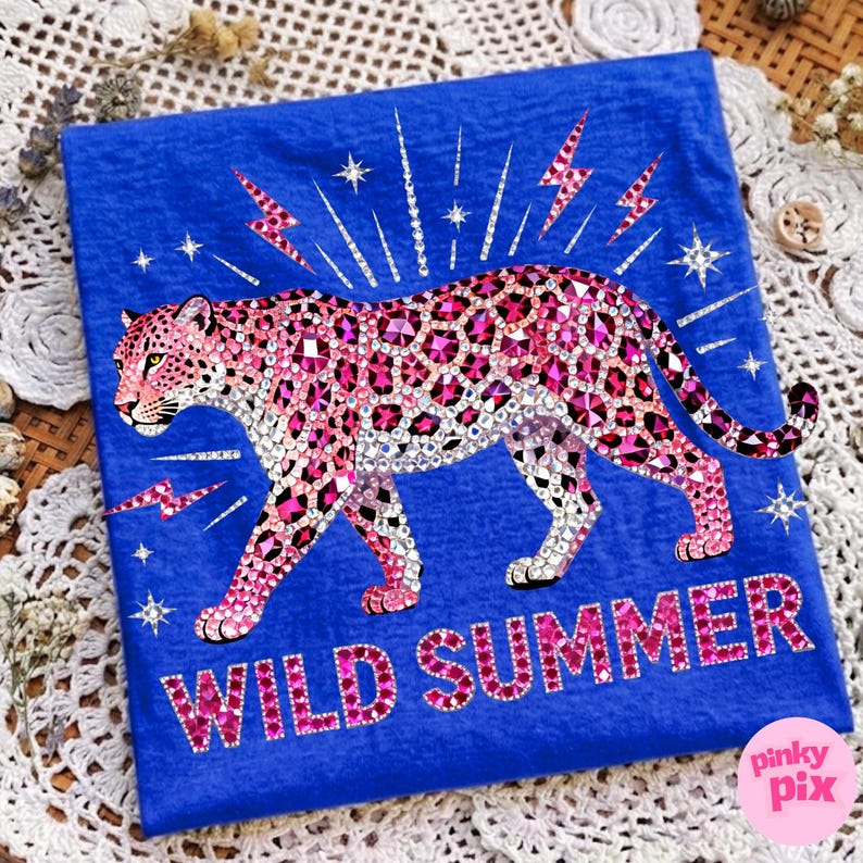 May include: A blue t-shirt featuring a pink leopard design with rhinestone accents. The leopard is surrounded by lightning bolts and stars. The words "WILD SUMMER" are displayed in pink rhinestones. The shirt has a Pinky Pix logo in the bottom right corner.