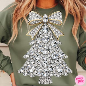 May include: A green sweatshirt featuring a sparkling Christmas tree design. The tree is adorned with numerous clear, faceted gems, and topped with a bow. The bow and tree base have gold accents. The design is centered on the front of the shirt.