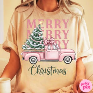 May include: A beige t-shirt featuring a pink vintage truck carrying a Christmas tree and a large bow. The words "MERRY CHRISTMAS" are printed in pink and green. The shirt is a festive holiday design.