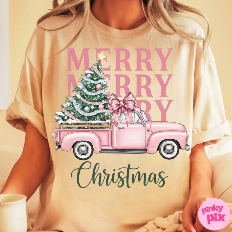 May include: A beige t-shirt featuring a pink vintage truck carrying a Christmas tree and a large bow. The words "MERRY CHRISTMAS" are printed in pink and green. The shirt is a festive holiday design.