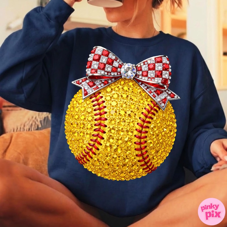 May include: Navy blue sweatshirt with a large, sparkling softball design. The softball is yellow with red stitching and a red and white checkered bow with a central diamond. The sweatshirt is a solid color.