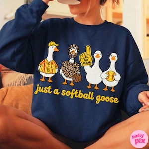 May include: Navy blue sweatshirt featuring cartoon geese in baseball attire. The geese are wearing a baseball uniform, leopard print, and holding a baseball and a "Go Team" sign. The text "just a softball goose" is in yellow.
