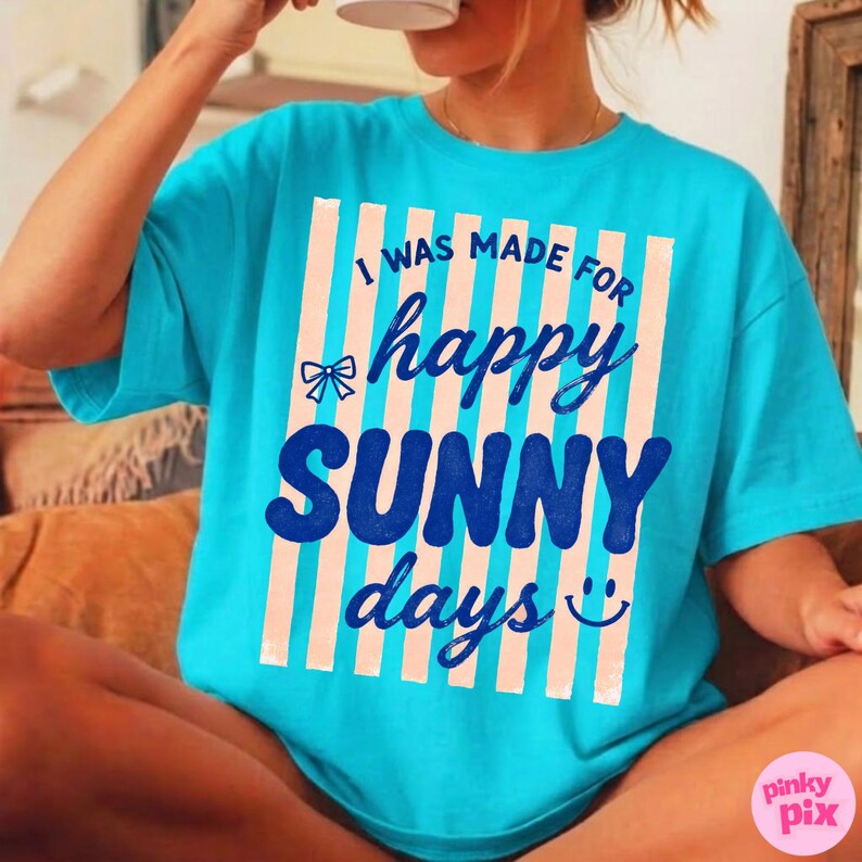 May include: A turquoise t-shirt with the text "I WAS MADE FOR happy SUNNY days" in a vertical striped design. The text is in a mix of fonts and colors, with a bow graphic and a smiling face. The shirt is a casual, relaxed fit.