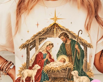 May include: A cream-colored t-shirt featuring a graphic of a nativity scene. The print shows a stable with a star above, Mary in a red robe, Joseph in a green robe, and a baby in a manger. Two lambs are present, one on each side of the scene.