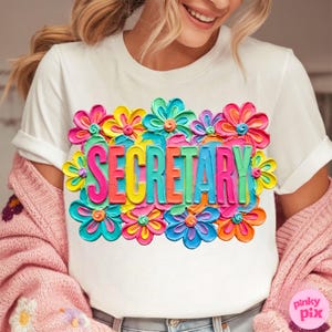 May include: White t-shirt with the word "SECRETARY" in colorful, 3D-style lettering, surrounded by vibrant, multi-colored flower graphics. The shirt is made of a soft material.