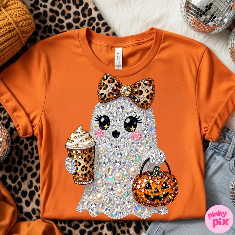 Może przedstawiać: Pomarańczowy t-shirt z grafiką ducha ozdobionego przezroczystymi i opalizującymi dżetami. Duch ma kokardę w panterkę, trzyma kubek do kawy w panterkę i niesie wiaderko w kształcie dyni z pomarańczowymi dżetami.
