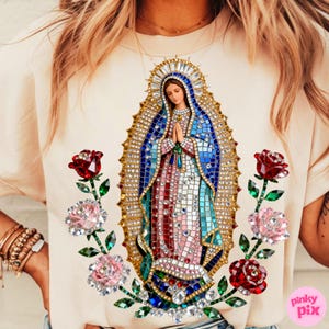May include: Cream-colored t-shirt featuring a mosaic-style image of the Virgin Mary, adorned with sparkling embellishments. The design includes a halo, blue and red robes, and is flanked by jeweled roses. The text "pinky pix" is visible.