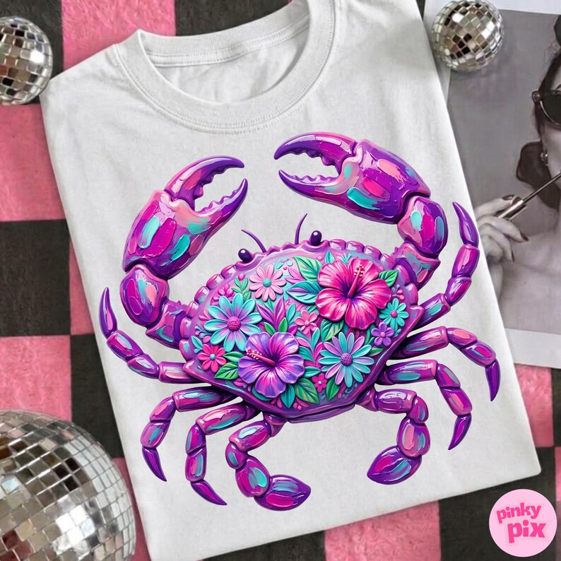 May include: White t-shirt featuring a vibrant crab design. The crab is rendered in shades of purple, pink, and teal, adorned with floral accents. The design is centered on the shirt, creating a bold and eye-catching graphic.