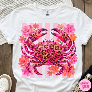 May include: White t-shirt featuring a vibrant pink crab with a leopard print design on its shell and claws. The crab is surrounded by pink and orange flowers, set against a pink striped background.