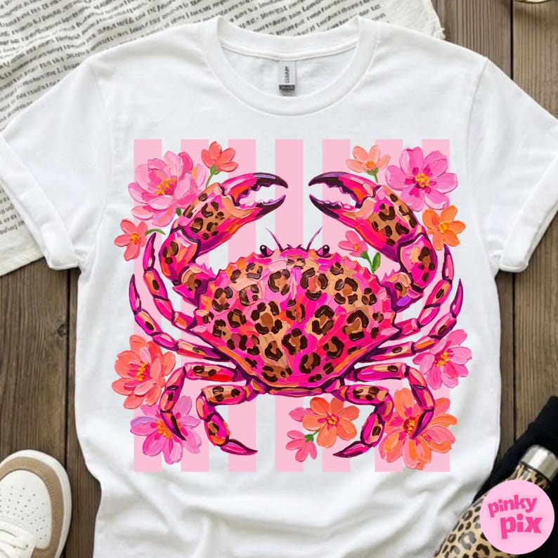 May include: White t-shirt featuring a vibrant pink crab with a leopard print design on its shell and claws. The crab is surrounded by pink and orange flowers, set against a pink striped background.