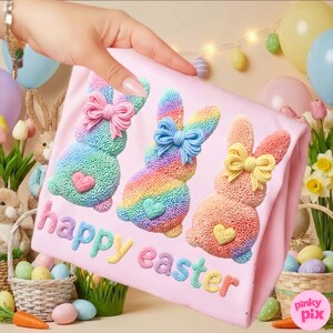 以下が含まれることがあります： ピンク色のイースターをテーマにしたシャツ。3匹のカラフルなウサギのデザインで、それぞれにリボンとハートが付いています。ウサギは虹色で、シャツには「happy easter」の文字がパステルカラーで刺繍されています。