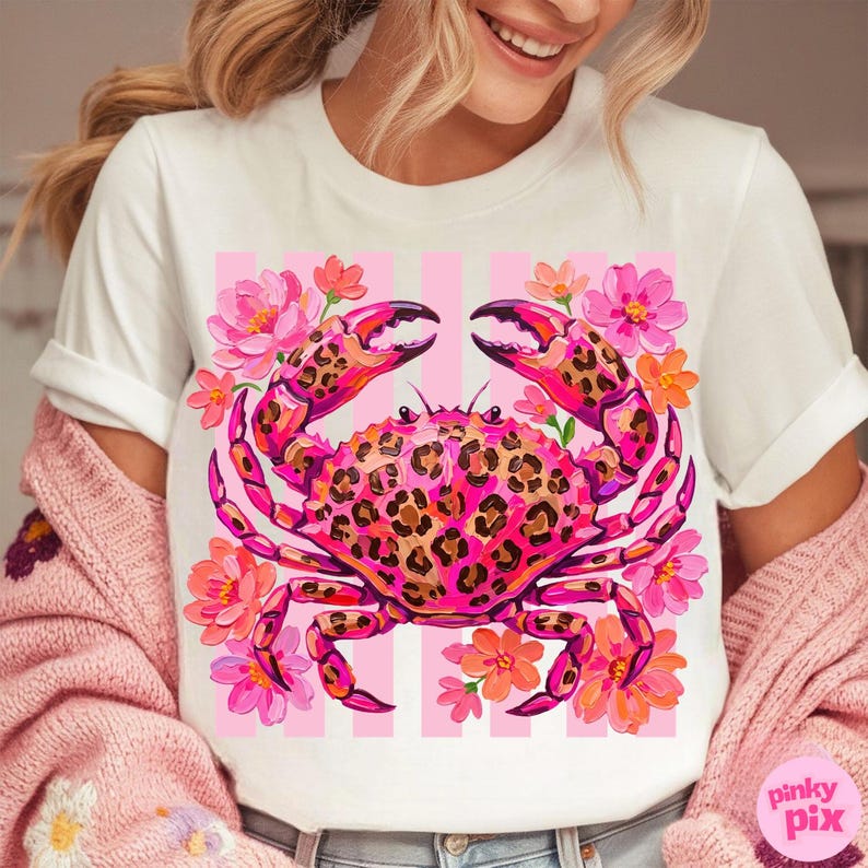 May include: White t-shirt featuring a pink crab with a leopard print design, surrounded by pink and orange flowers. The crab is set against a pink and white striped background. The shirt has rolled-up sleeves.