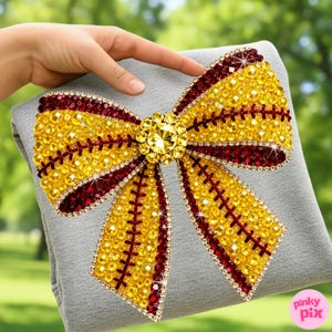 May include: A large, decorative bow with a softball design. The bow is embellished with yellow and burgundy rhinestones, and a central yellow jewel. Red stitching details add to the sparkling, eye-catching accessory.