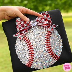 May include: A baseball-themed decorative item featuring a baseball covered in sparkling crystals and red stitching. A red and white crystal bow adorns the top. The baseball is set on a black surface.