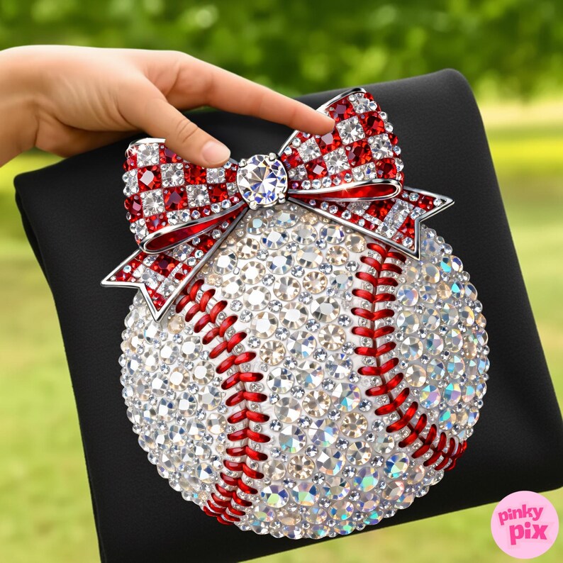 May include: A baseball-themed decorative item featuring a baseball covered in sparkling crystals and red stitching. A red and white crystal bow adorns the top. The baseball is set on a black surface.