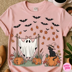 May include: Pink t-shirt with a Halloween-themed graphic. The design features a ghost hanging on a clothesline, pumpkins, a black cat wearing a witch hat, bats, and autumn leaves. The shirt is made of a soft material.