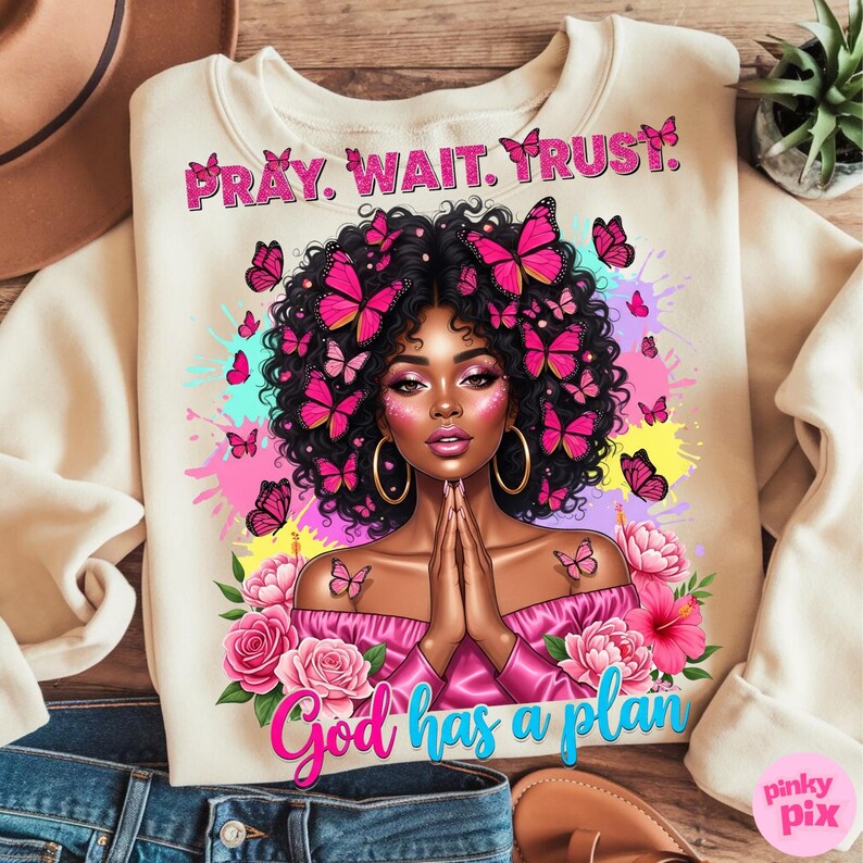May include: Cream-coloured sweatshirt with a graphic of a woman adorned with butterflies. The text "Pray. Wait. Trust." is above the image, and "God has a plan" is below. The design incorporates pink and magenta hues, creating a vibrant aesthetic.