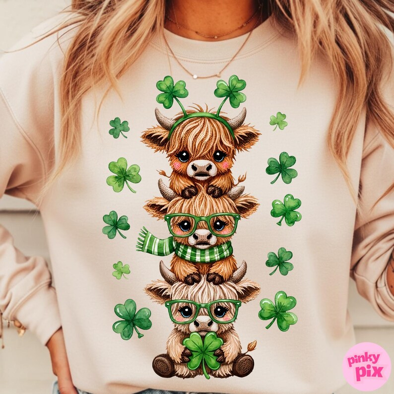 May include: A light beige sweatshirt featuring a stack of three cartoon Highland cows. The cows are brown with green accessories, including glasses, a scarf, and shamrock headbands. Green shamrocks are scattered around the cows.