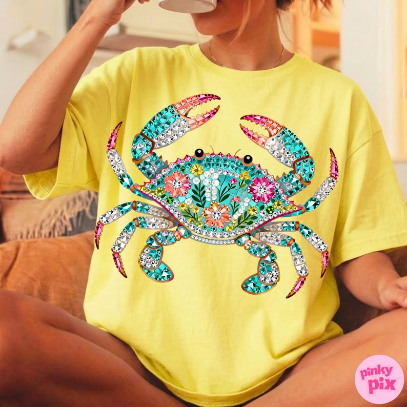 May include: Yellow t-shirt featuring a large, ornate crab design. The crab is decorated with turquoise, pink, and clear jewel-like accents, and floral patterns. The shirt has short sleeves and a relaxed fit.