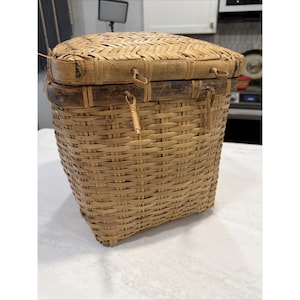 May include: A square-shaped woven bamboo basket featuring a natural tan finish and a fitted lid. The basket includes a reinforced rim with two small loops on the front and two on the lid, connected by thin, flexible bamboo strips for closure.