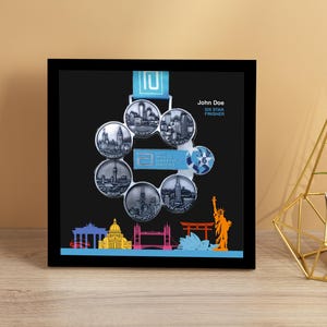 May include: A framed black display featuring six circular medals with cityscapes, connected by a blue ribbon. The frame includes the name "John Doe" and "Six Star Finisher". A gold geometric plant holder and a small potted plant are to the right.
