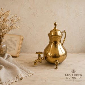 May include: A polished brass beverage dispenser featuring a rounded body, a pointed lid, a curved handle, and a small spigot at the base. The vessel stands on three short legs and has a metallic gold finish that reflects light across its smooth surface.