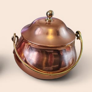 Peut inclure: Une petite marmite de couleur cuivre avec un couvercle et une poignée. La marmite a un corps arrondi et un couvercle avec un petit bouton rond. La poignée est de couleur or et s'incurve sur les côtés. La marmite est posée sur une surface claire.