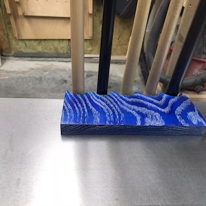 May include: A blue and white wooden object with a wave-like design. The object is rectangular and sits on a metal surface. The wood grain is visible, and the object is in a workshop setting.
