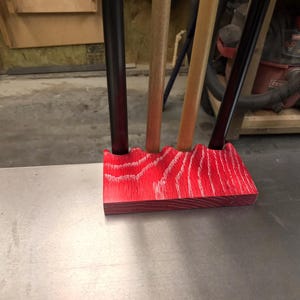 May include: A red wooden tool holder with a wavy top, holding several tools with black and wooden handles. The holder is made of red-stained wood with a visible grain pattern, sitting on a metal surface.